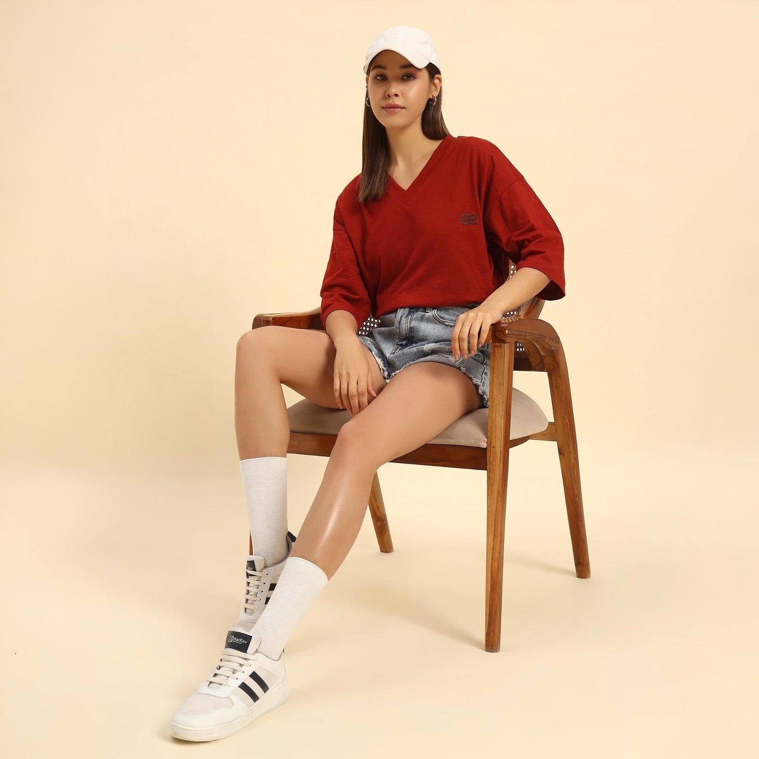 Woman in red V-neck shirt and denim shorts sitting on a chair, wearing white cap and sneakers, aeternum fashion