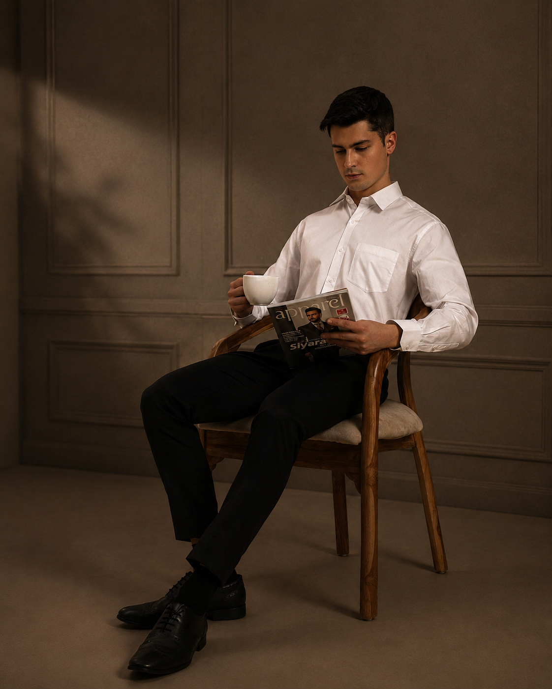 Man in a premium white shirt from aeternum sitting on a wooden chair, reading a magazine, and holding a mug.